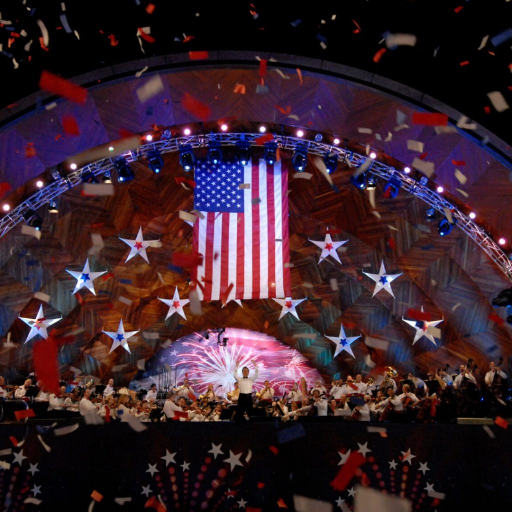 This photo is from a Fourth of July celebration at the Hatch Memorial Shell on the Charles River Esplanade. The photo shows red white and blue confetti falling from the ceiling, with decorative stars hanging from the tall ceilings, as well as an American flag. There are people in the crowd and there is a stage with a band performing at it. Photo courtesy of bso.org. 