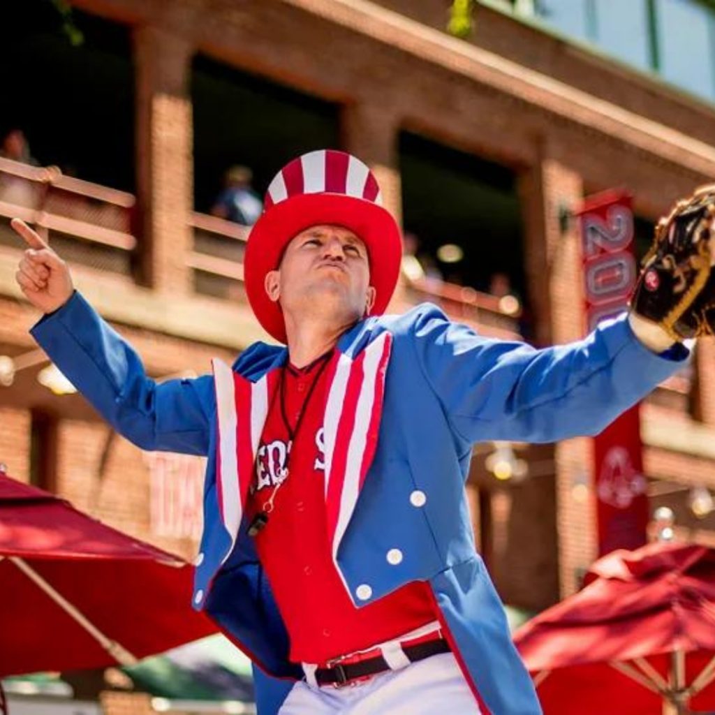 This is a photo from the Fourth of July at Fenway Park. There is a man standing out side of Fenway Park, wearing patriotic red white and blue gear. He is wearing a Red Sox jersey underneath, with a brown baseball glove on his hands. Picture credits: MLBlogs.com