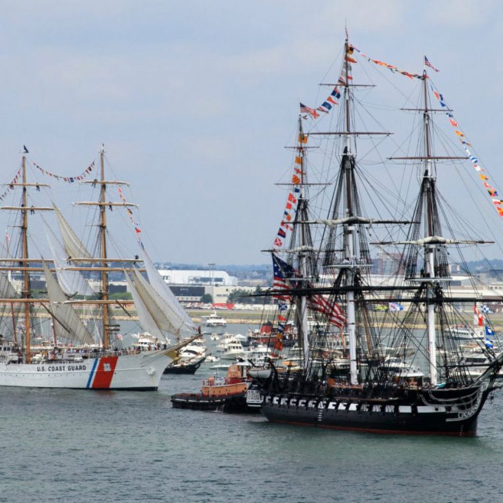 This photo shows two ships and a few smaller boats that are sitting in Boston Harbor. The water is a mix of blues and there is over cast in the sky. One of the ships is historic looking and the other is a Coast Guard ship. Picture credits: libertyfleet.com
