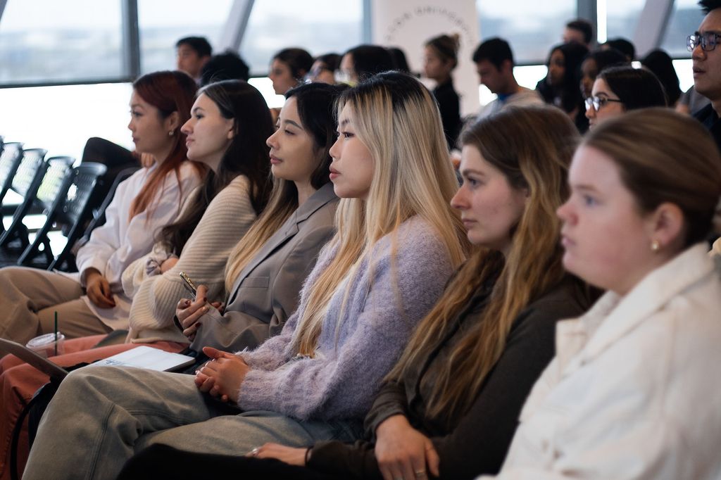 Photo: Students sit in rows of chairs as they face the front of a brightly lit room.