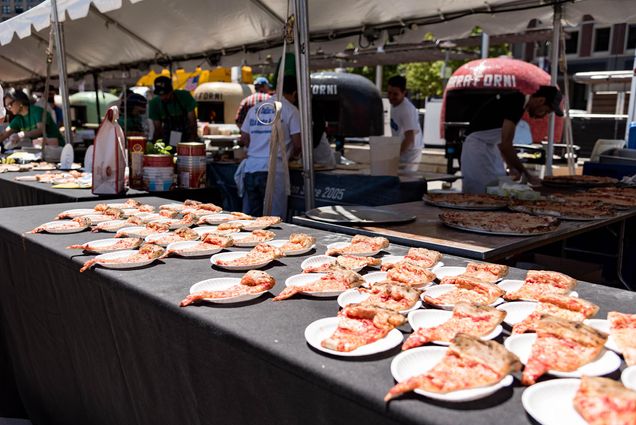 Photo: A long table with a black cloth with multiple paper plates featuring a large slice of cheese pizza. The photo was taken outside, the day sunny and bright.