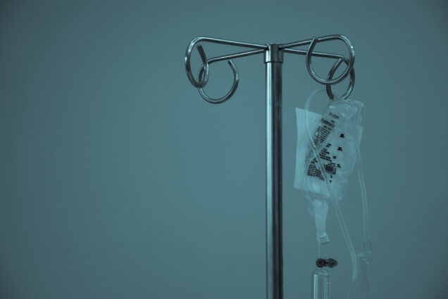 Photo: A dark blue background with a metal IV holder with an IV bag strung up on one of the hooks.