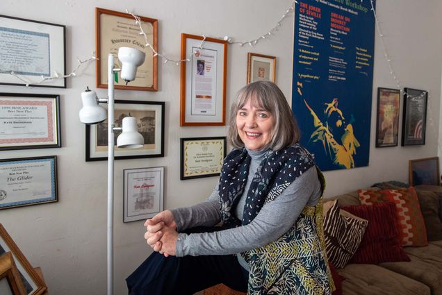 Photo of Kate Snodgrass, a middle aged White woman with a gray bob, who wears a dark blue scarf with white pattern. She sits on the corner of a couch, presumably in her office, where framed awards and playbills hang on the wall behind her.