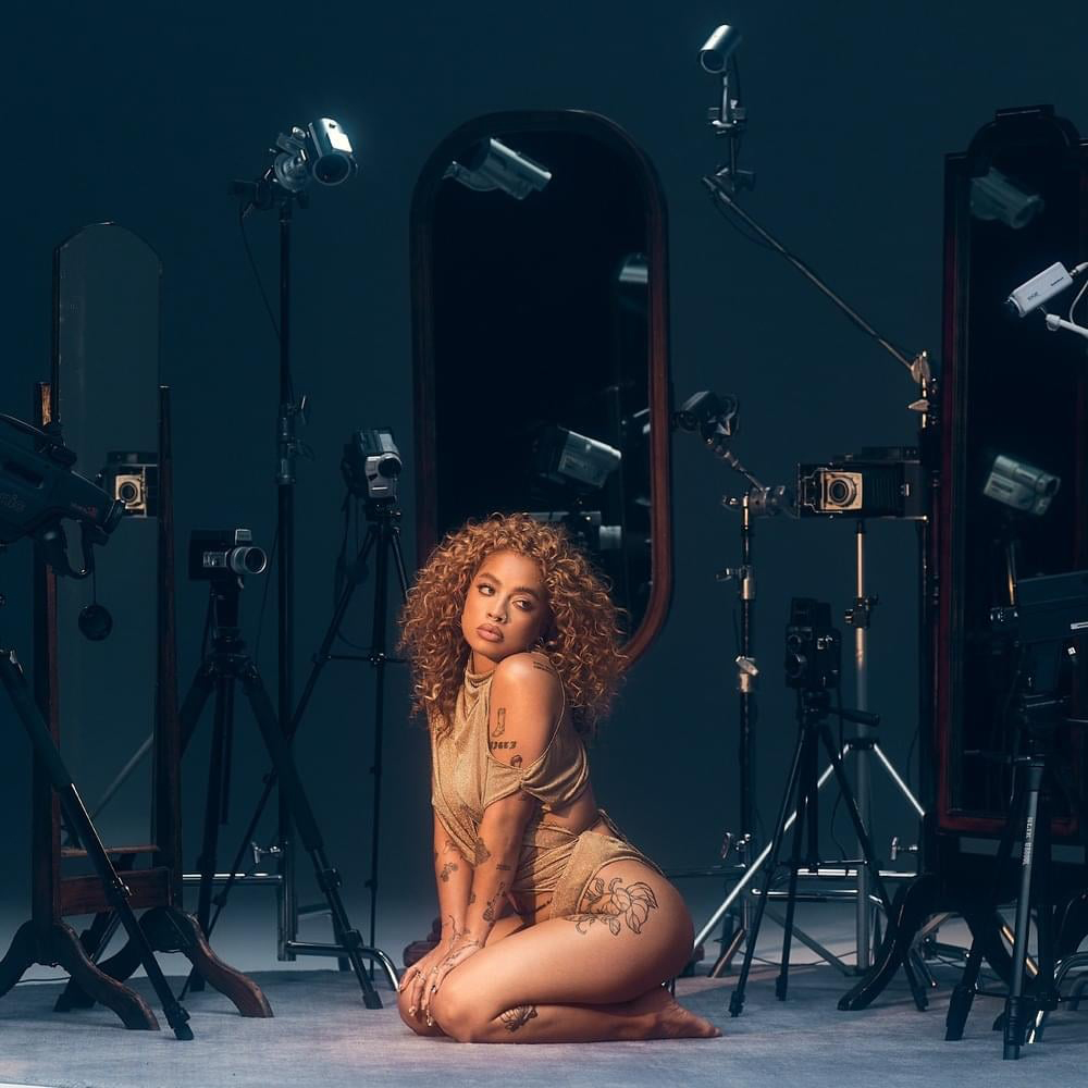 Photo: A Black womanm Kiana Lede, sitting with her legs under her and her hands on her knees with her face over her shoulder. Various cameras surround her and point to where she is seated.