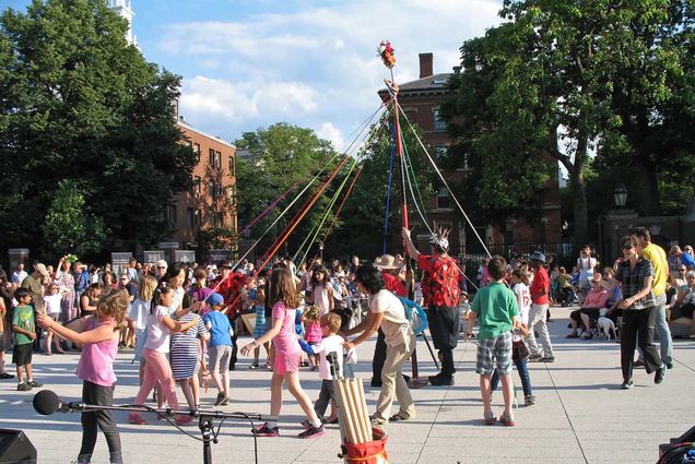 Photo: A large crowd walks around a arial type structure with large ribbons attached to a tall pole. The ribbons are a various of different colors. The crowd is moving to the left. The day is bright.