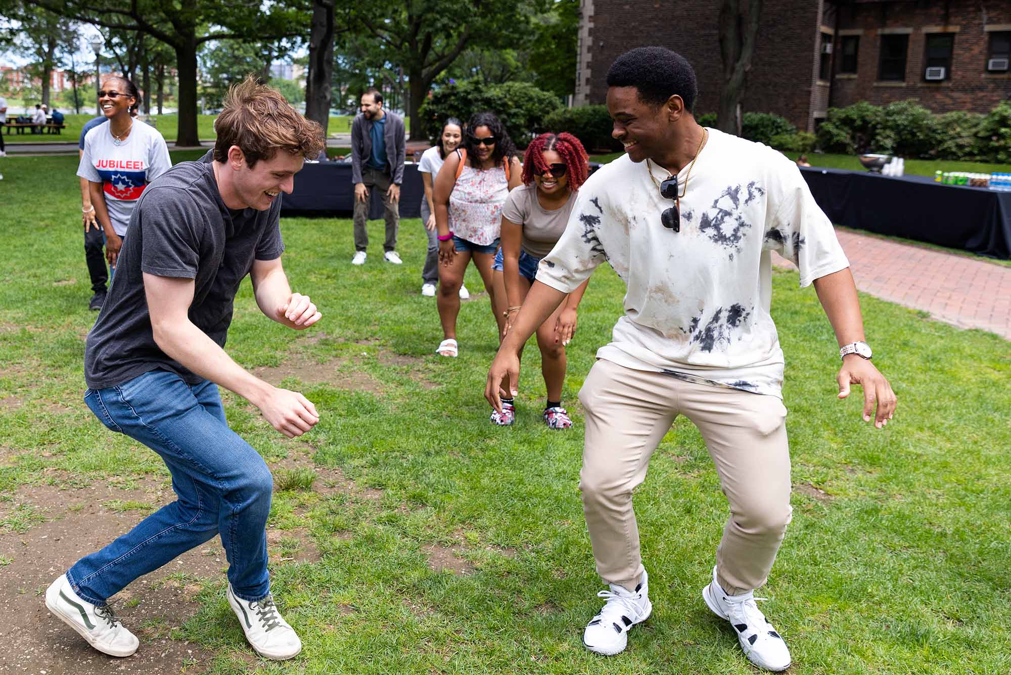 Photo: A few individuals dance to the Cha Cha slide of a grassy field. Two of the individuals in the forefront of the photo are (from left) a white man with a dark tshirt and jeans. He has floppy brown hair. On his right, his roommate, a Black man with a white button up and lighter pants smiles and dances with him.