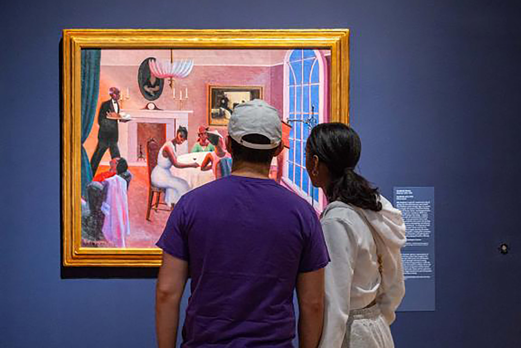 Photo: A photo of two individuals looking at a painting at the MFA. The wall is blue, the painting a pink haze, and the first individual on the left is wearing a purple shirt and a white baseball cap while on the right, an all white outfit with a pony-tail.