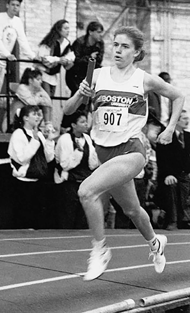 Black and white photo of Jennifer Lanctot O'Neill (Questrom'91), BU's first three-time female All-American, running with a relay stick. She wears a white tank top and shorts and has the number 907. People behind her can be seen cheering others in the race.