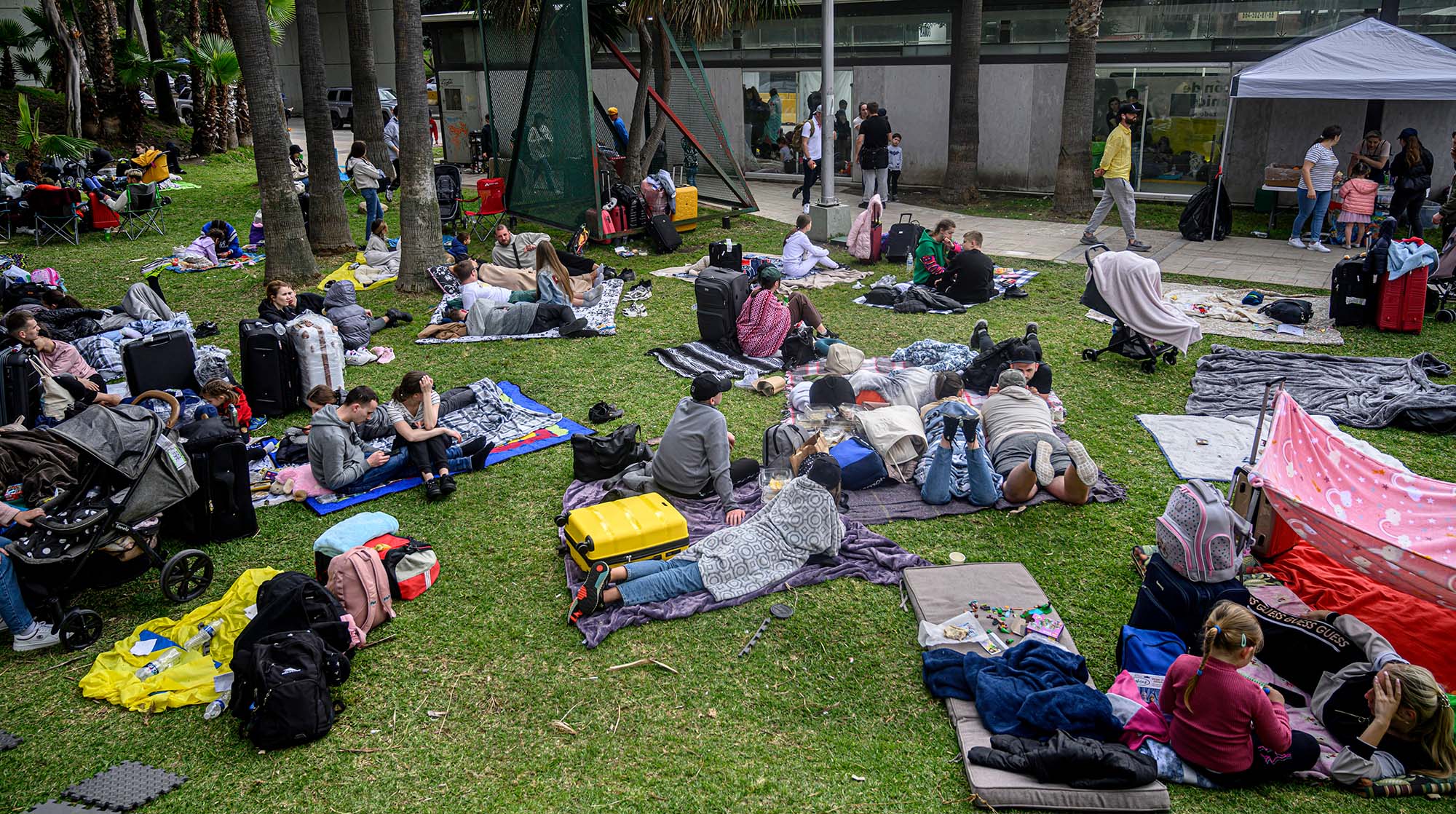 Photo of Ukrainian refugees awaiting entry into the US. They sit and lay on blankets on a patch of grass. Many wearing sweatshirts and jeans and look away from the camera towards a gray tent and wall. The refugees sit amongst their suitcases and there is a mix of families and groups of young people.