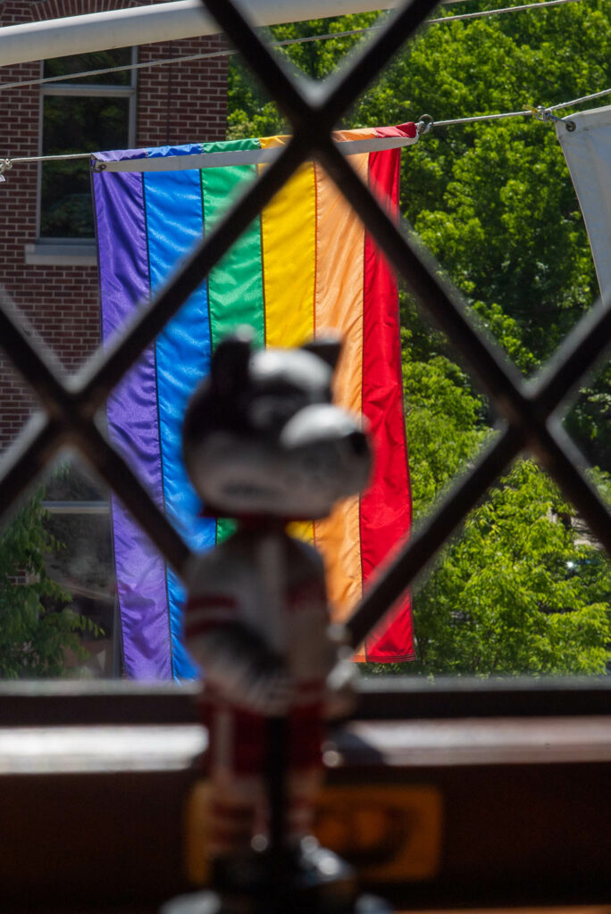 Photo: A window with bars on it with a Pride Flag hanging outside and a bobble head of BU's mascot, a terrier.