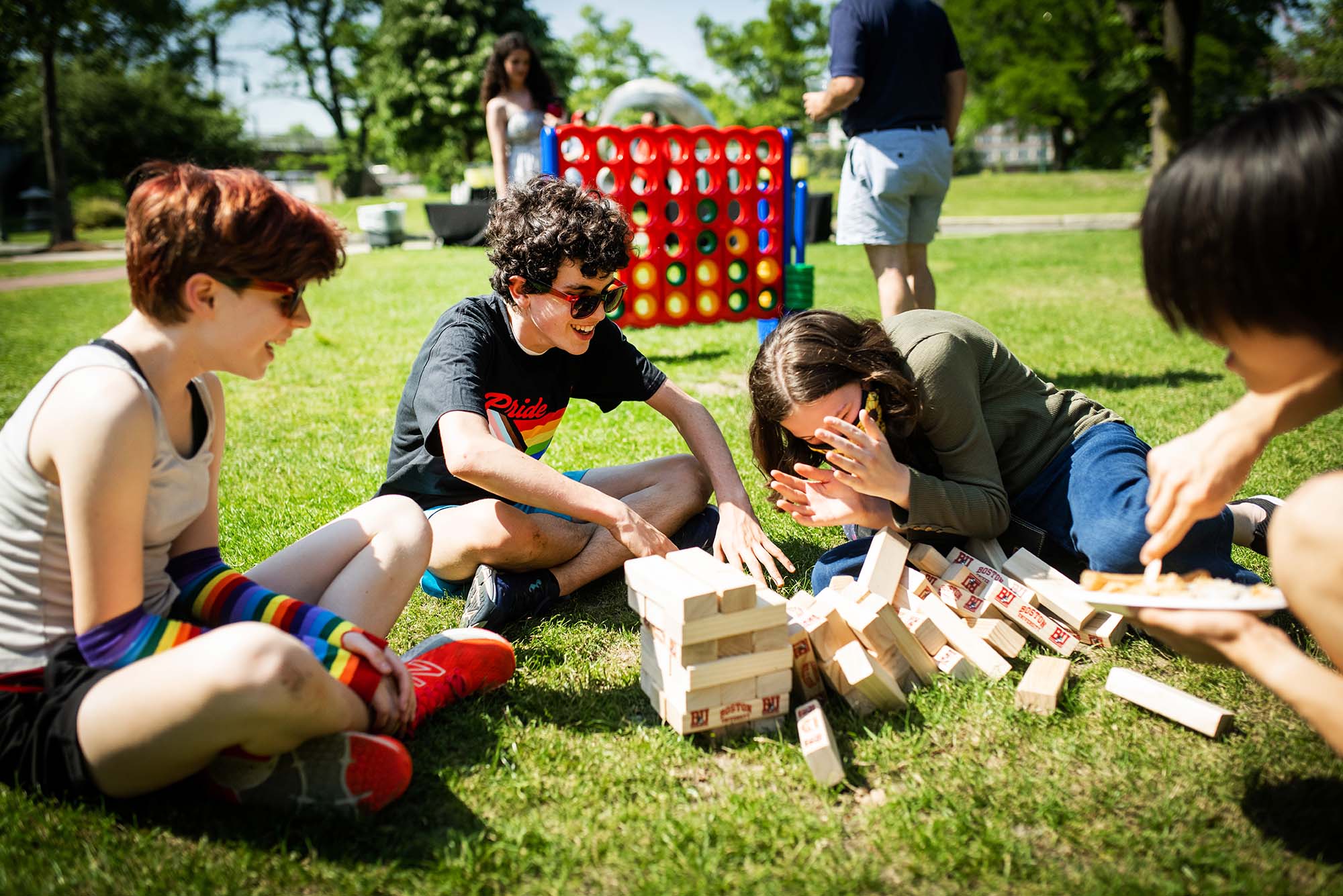 Photo: Four BU Academy students (left to right), Coco Mueller, a white individual with short brown hair and rainbow arm-warmers; Owen Bergstein, a white individual with a black t-shirt and glasses; Kaela Harrison, covering their face and wearing a long-sleeve gray shirt with jeans; and Andrew Chao, in the far right-most part of the frame sit on grass a play Jenga, a wooden block game, at an outdoor BBQ celebrating Pride Month. The day is bright and sunny.