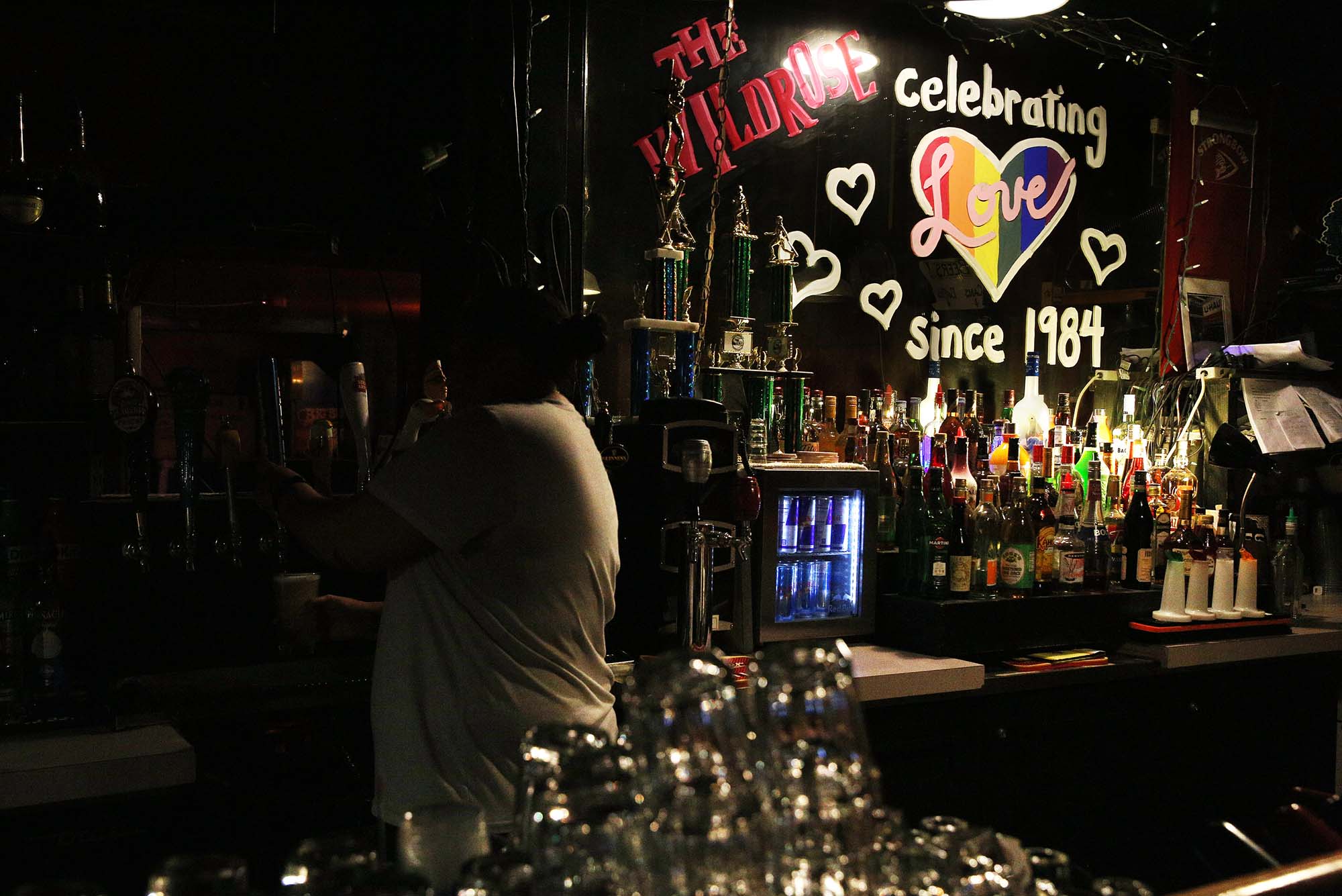 Photo of one of the oldest lesbian bars in the country, Wildrose in Capitol Hill taken Sept. 14, 2016. The bar is dimly lit. In the photo, a bartender in a white shirt, with their back to the camera, fills up a bar at a tap. Behind the bar, a mirror is decorated with window paint that reads "The Wildrose, celebrating Love since 1984." The word "love" is inside a rainbow heart and smaller white hearts encircle the larger heart.