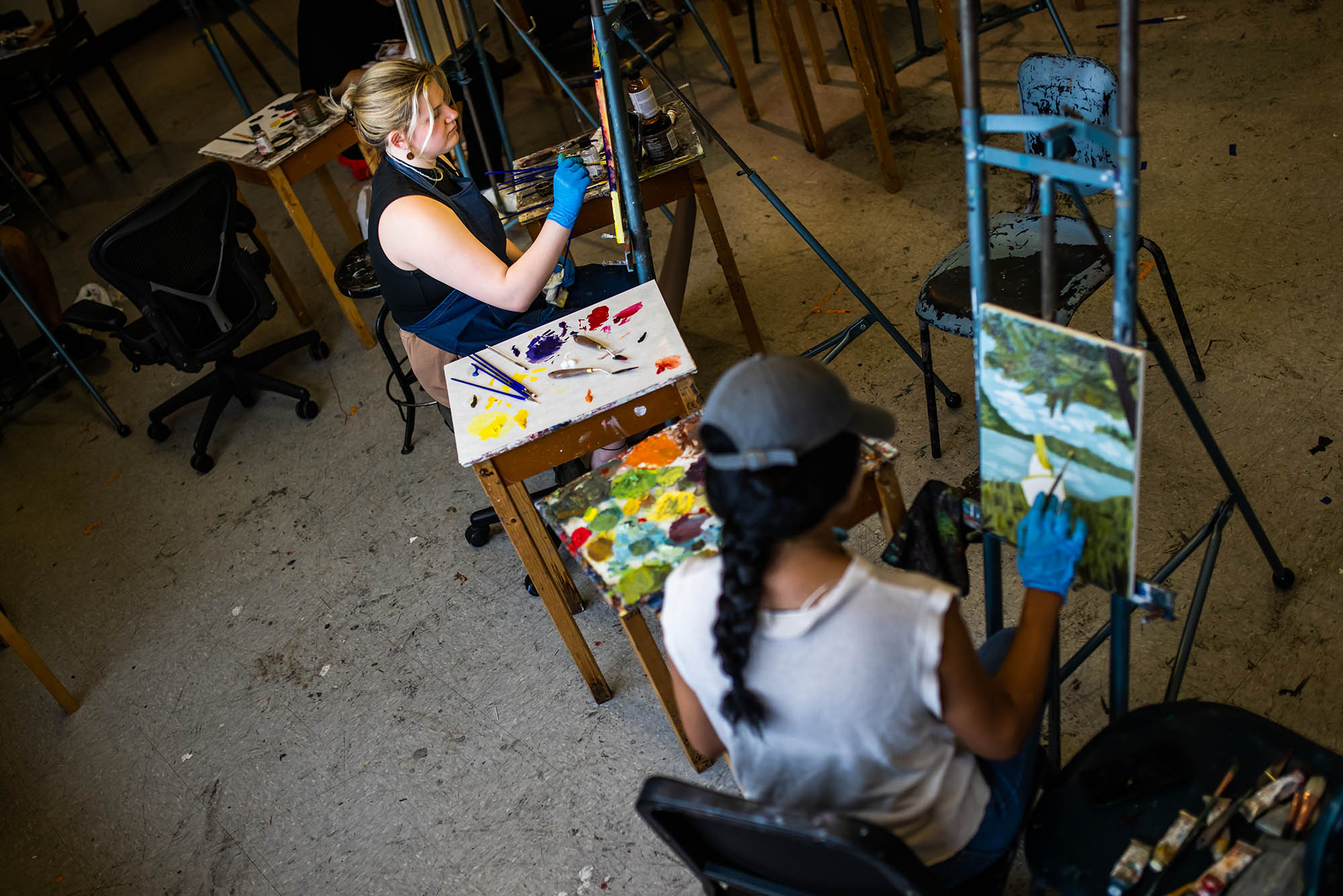 Photo: Overhead shot of two young women working on paintings. The white woman on the left wears a black sleeveless shirt as she works and the perosn on the rioght wears a white tank top and grey cap as they work on a nature scene.
