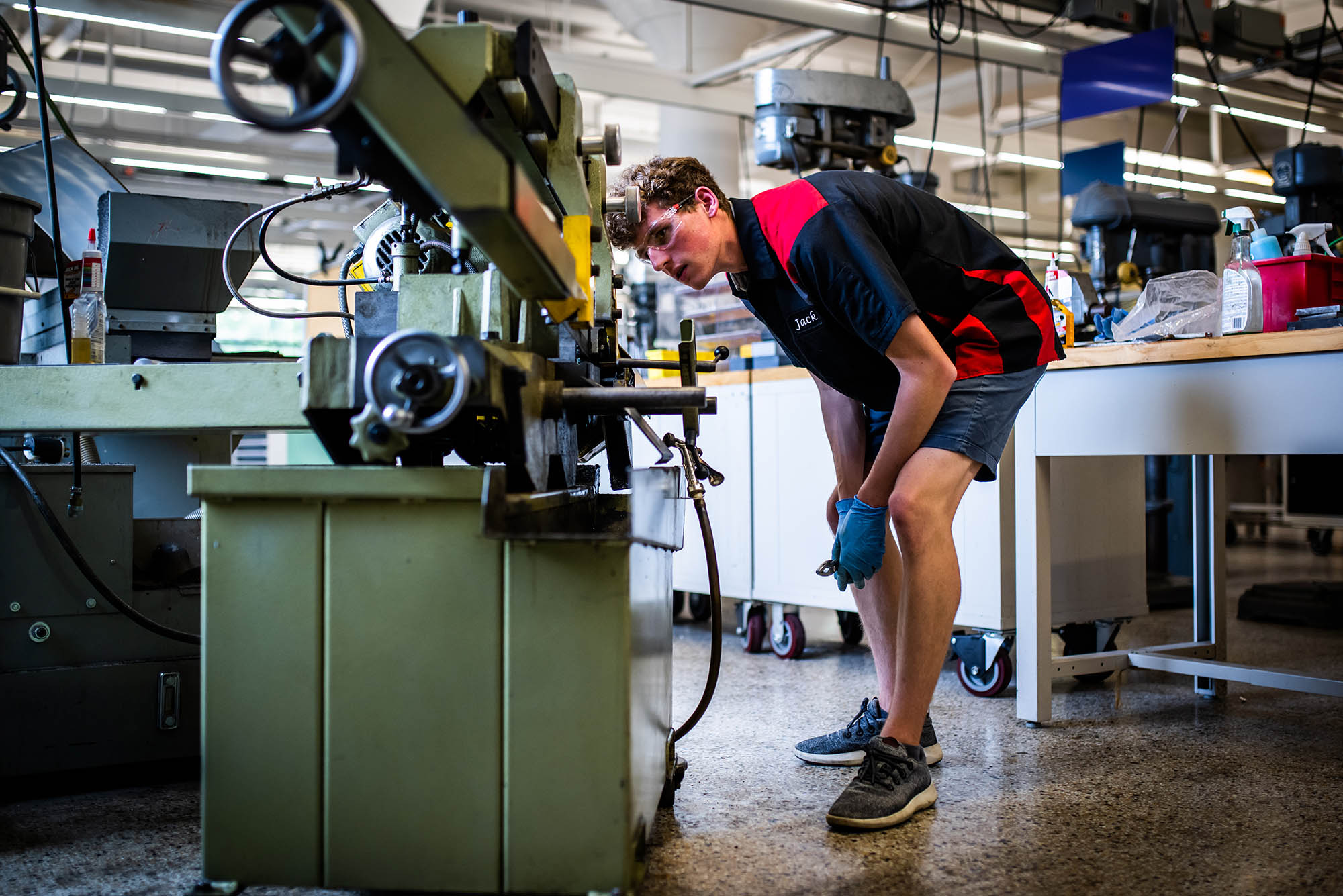 Photo: A young white man wearing clear plastic safety glasses, a red and black shirt with the name "Jack" printed on it, black shorts, and blue medical gloves bends and looks at a large band saw in front of him. Behind him, other mechanical items in a lab can be seen.