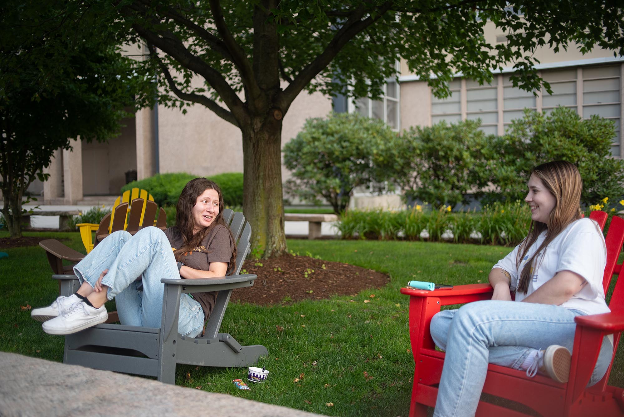 Photo: Two young women sit in large wooden grey and red lawn chairs. They look towards each other as they lounge and chat.