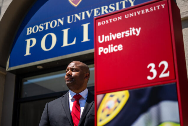 Photo: Robert Lowe, a Black man wearing a black suit, white collared shirt, and red ties, poses in front of the Boston University Police station. Behind him, the station can be sign with large blue sign that reads "Boston University Police". To his right, a red signpost reads "University Police 3"