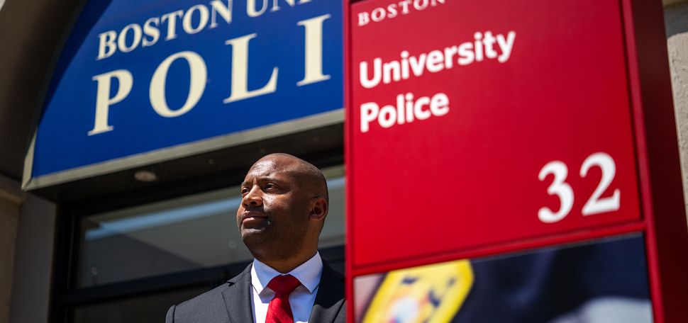 Photo: Robert Lowe, a Black man wearing a black suit, white collared shirt, and red ties, poses in front of the Boston University Police station. Behind him, the station can be sign with large blue sign that reads "Boston University Police". To his right, a red signpost reads "University Police 3"