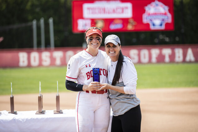 Photo: Caitlin Coker, a young white woman wearing a red Bu cap and a white and red BU softball uniform smiles and poses with a small, blue glass trophy. On her right, head coach softball coach Ashley Waters, a white woman with wearing a white and grey BU shirt and grey cap, smiles and poses with her. They both stand in front of a softball field.