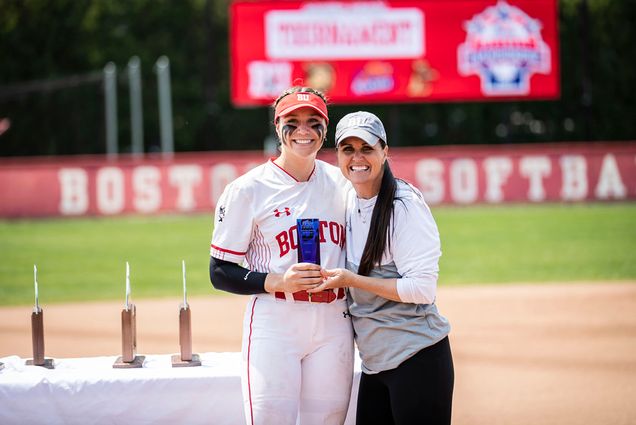 Photo: Caitlin Coker, a young white woman wearing a red Bu cap and a white and red BU softball uniform smiles and poses with a small, blue glass trophy. On her right, head coach softball coach Ashley Waters, a white woman with wearing a white and grey BU shirt and grey cap, smiles and poses with her. They both stand in front of a softball field.