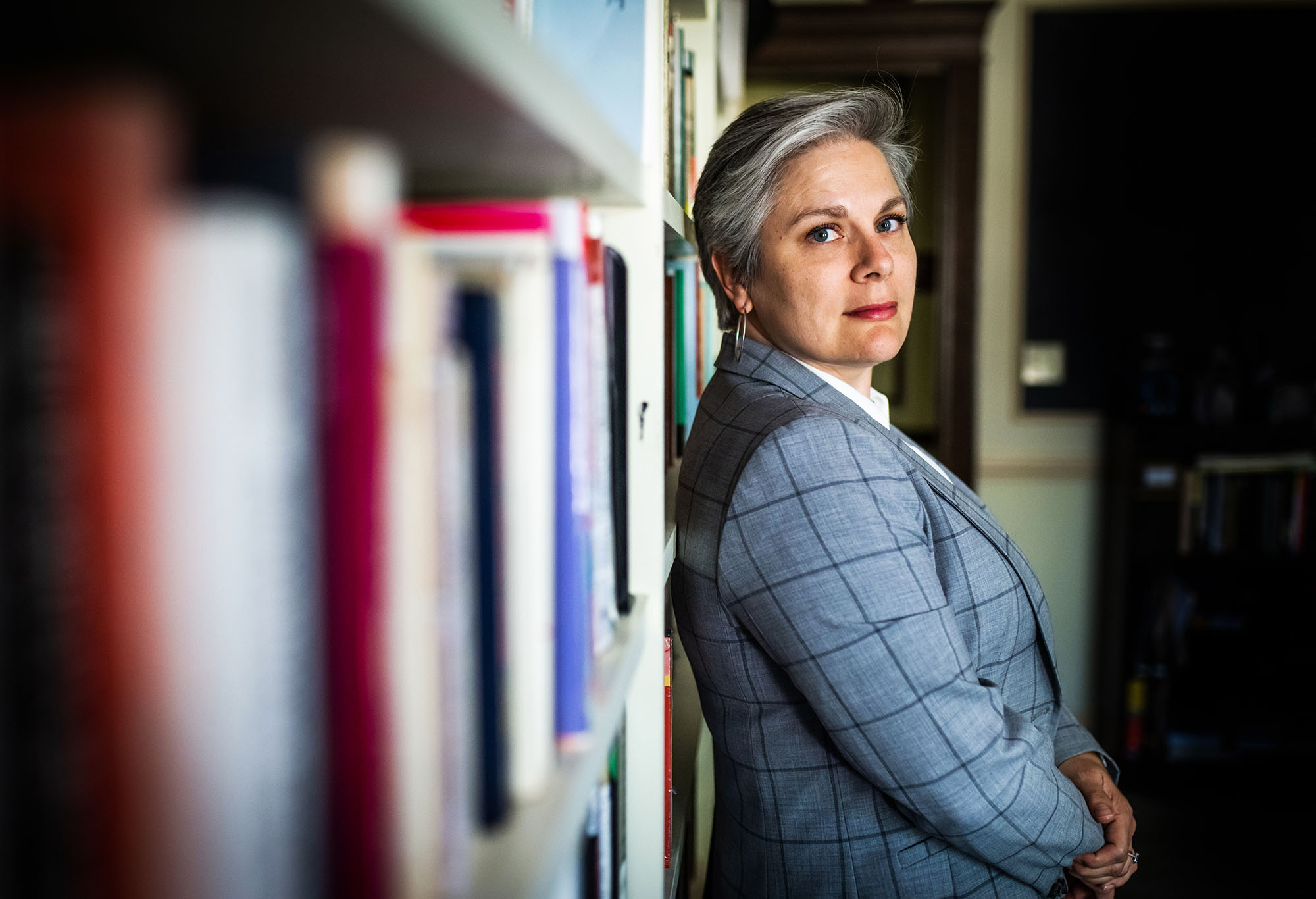 Photo: Cari Babitzke, a white woman with short silver-grey hair and wearing a blue-grey blazer and white collared shirt, leans her back against a bookcase and looks to the camera on her right.