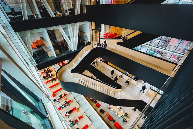 Photo: Overhead shot of students walking and gathering in and around a large set of black and wood butterfly stairs.