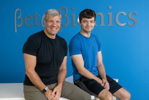 Photo: Beta Bionic’s Ed Damiano (left) with his son, David Damiano. A white man silver grey hair and wearing a black shirt and grey pants sits and poses next to a younger man with dark brown hair who is wearing a blue shirt and black pants. Both smile and pose in fornt of a blue wall. Silver letters on wall read "Beta Bionic".