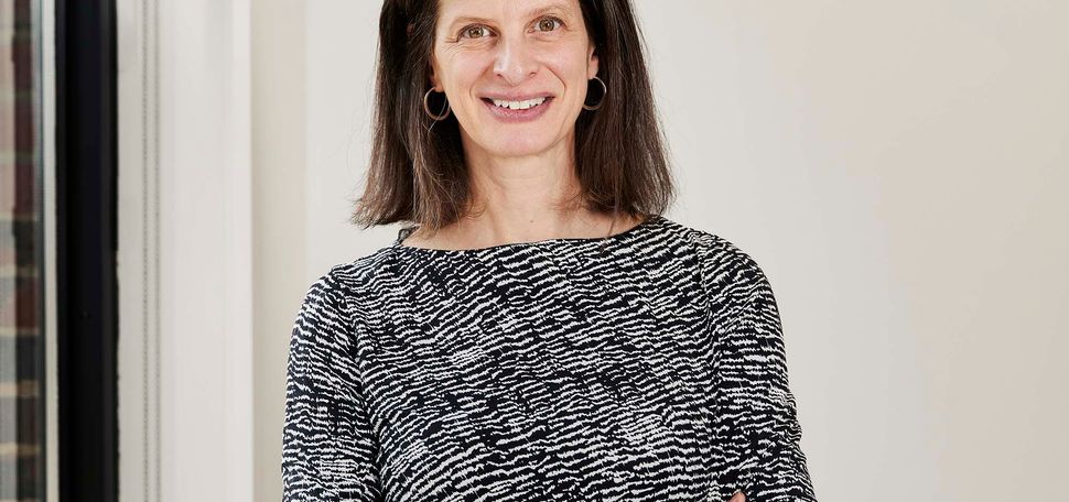 Photo: Dr. Emily Rothman, a white woman with shoulder-length brown hair and wearing a black and white half-sleeved dress, stands and poses with arms crossed. She smiles as she stands next to a large window in a brightly lit space.