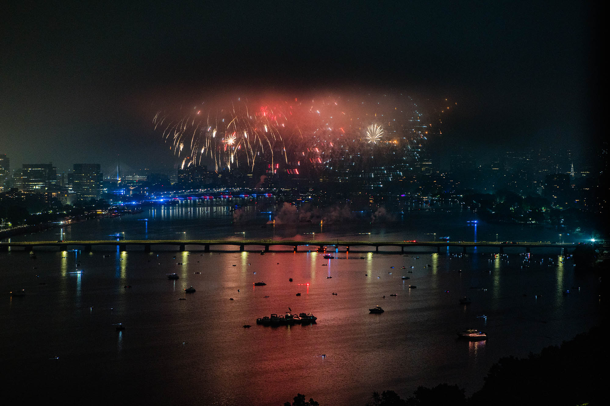 Photo: 4th of July fireworks are shown going off on a dark night above the Charles river. Red, white, and orange fireworks light up the night sky and show the Boston skyline.