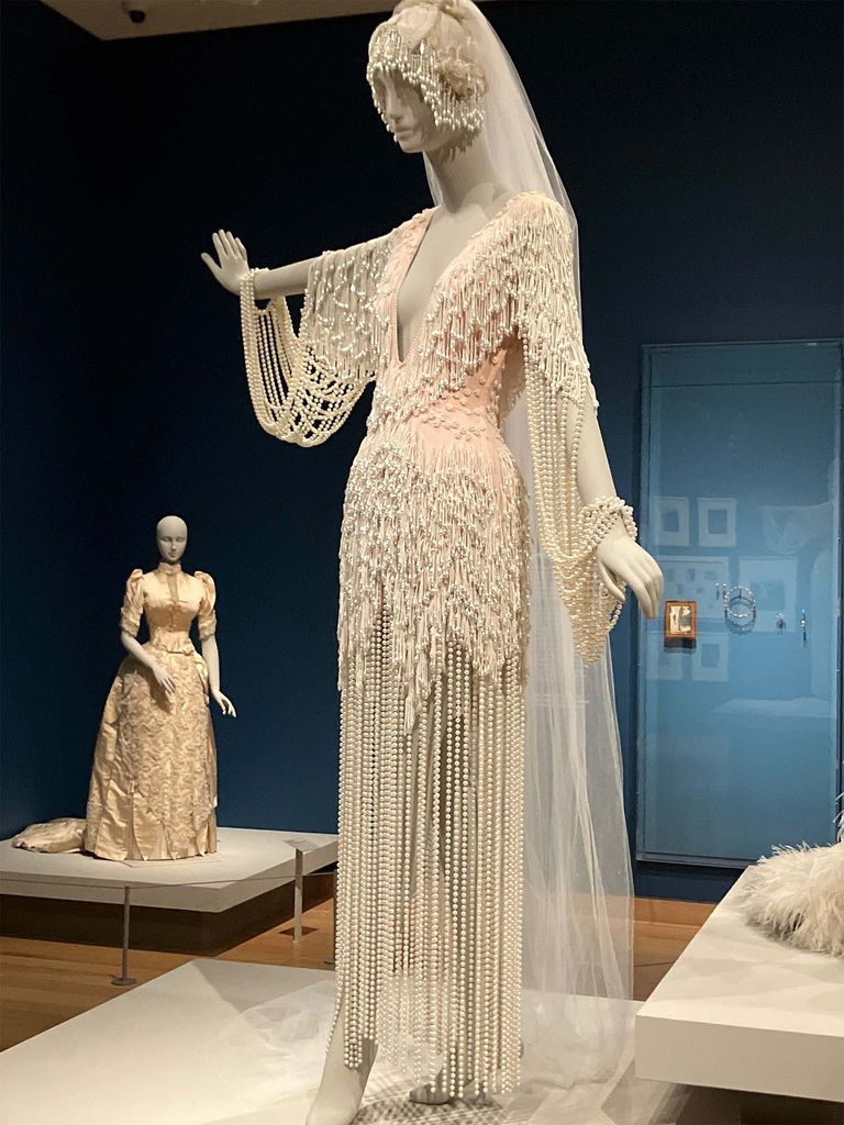 Photo: Two mannequins wearing intricate white wedding dresses with small plaques on the wall behind them