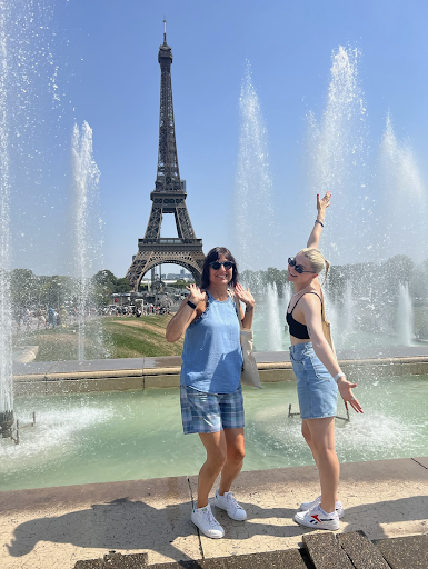 This is a photo of Boston University student Fia McCarty and her mother in front of the Eiffel Tower in Paris, France.  They are in front of the fountains and you can see the tower in the background.  Mom has brown hair and sunglasses and wears blue shorts and a blue shirt.  She has her hands up in the air and a smile on her face.  Fia looks down on her mother in denim shorts and a black tank top.  She opens her arms and smiles at the camera.  She has blonde hair and sunglasses.  The picture is bright and it looks sunny. 