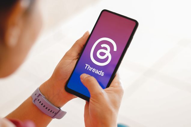 Photo: The app Threads from Meta is displayed on a mobile phone. A person holds up a phone that shows the large Threads logo and the word "Threads" on a gradient pink and purple background.