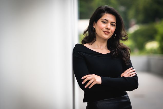 Photo: Nikta Khani, a young woman with long, curled black hair and wearing an off the shoulder black long-sleeved top and black pants, smiles and leans against the exterior of a white building. She leans with arms crossed.