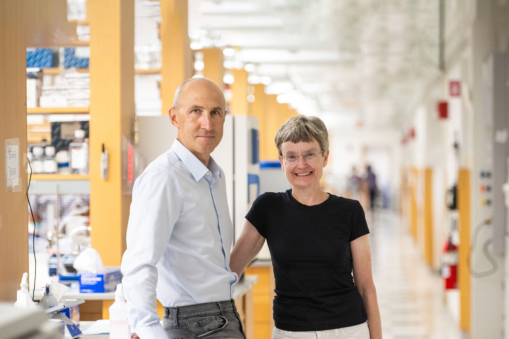 Photo: Ruslan Afasizhev (left), a tall whie man with a balding, shaved grey hair and wearing a light blue collared shirt and dark grey jeans, and Inna Afasizheva, a white woman with short grey-brown hair and wearing a black short-sleeved shirt and white jeans, pose in a brightly lit lab. Ruslan leans slightly back against a lab worktop while Inna poses with one hand one the same worktop behind Ruslan.