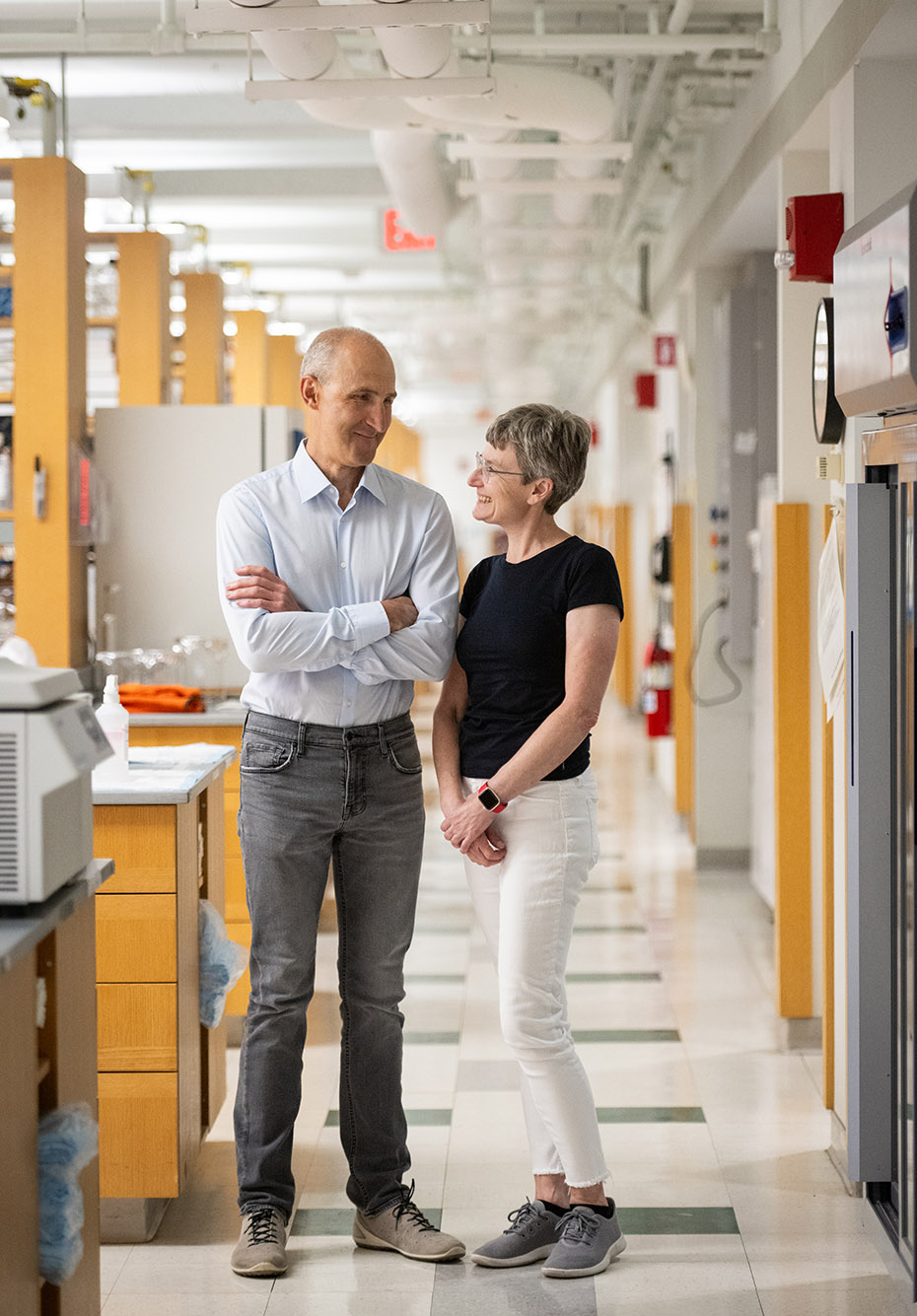 Photo: Ruslan Afasizhev (left), a tall whie man with a balding, shaved grey hair and wearing a light blue collared shirt and dark grey jeans, and Inna Afasizheva, a white woman with short grey-brown hair and wearing a black short-sleeved shirt and white jeans, pose in a brightly lit lab. Ruslan stands with arms crossed while Inna stands with hands clasped in front of her. Both smile and look to each other lovingly.