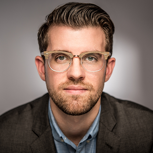 Photo: Carl Streed, a white man with brown hair and wearing glasses, a light blue collared shirt, and dark grey blazer, is shown posing in front of a grey background.