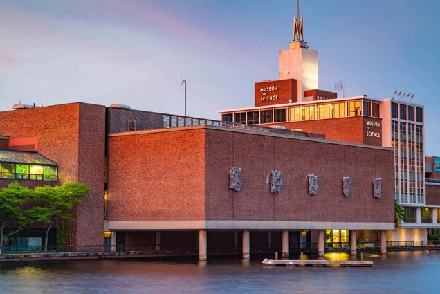 Photo: A landscape photo of The Museum of Science at dawn. The sky is blue with soft purples and pink and the sun is golden, refelcting off the top of the building.