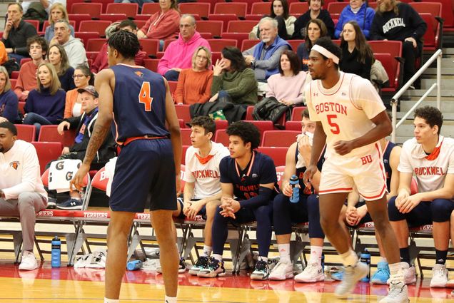 Photo: Walter Whyte, a tall, young Black man wearing a white and red BU Men's basketball uniform and white headband, moves across the court. A Bucknell athlete in a blue and orange uniform faces away from the camera toward center court. Player sit on the bucknell player bench behind them. A few people sit in the red seats in the stands in the background.