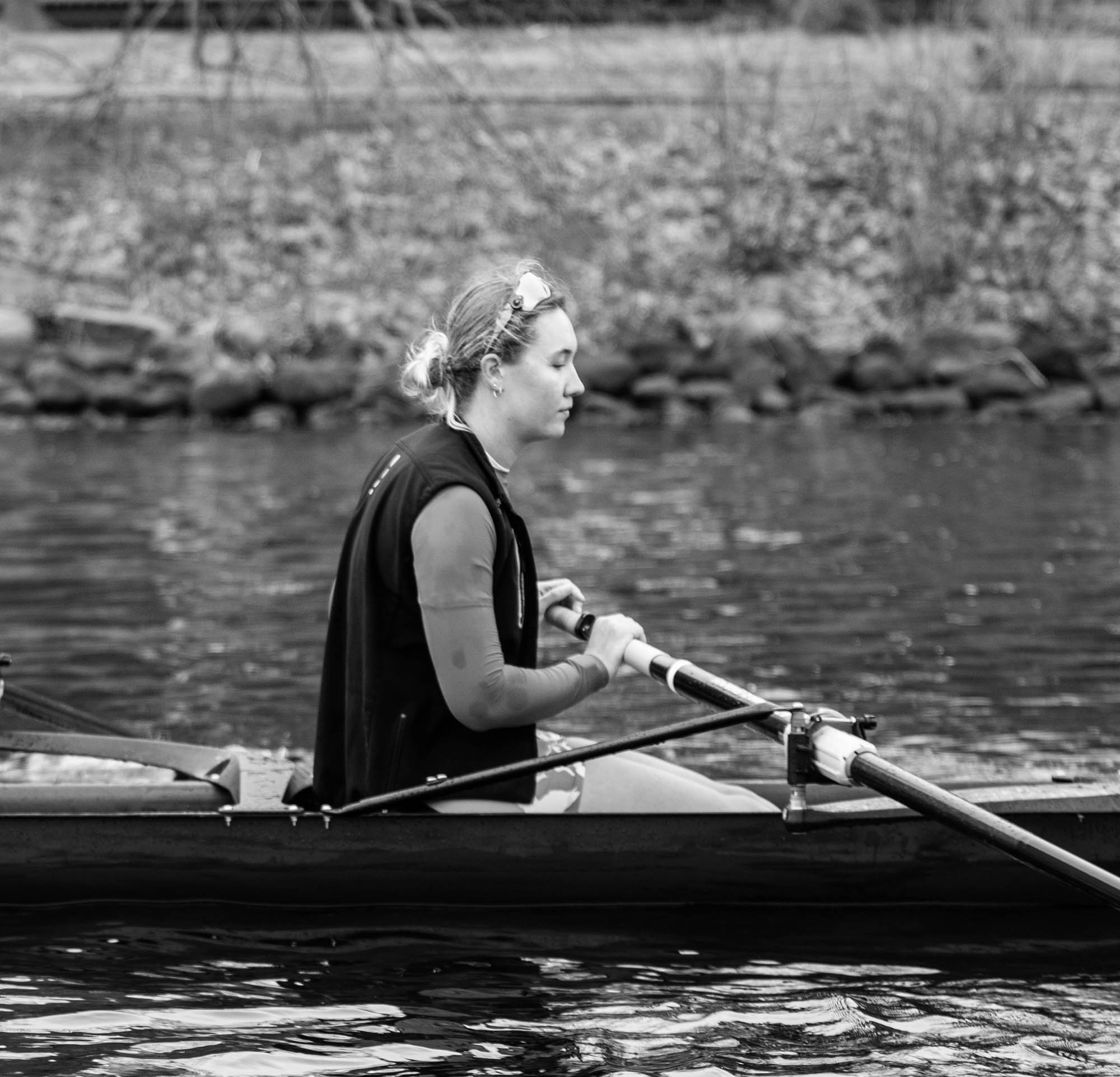 Photo: Black and white photos of Kat Robbins rowing with her team on the water. A young white woman with hair tied back in a bun and wearing a long-sleeved shirt, black vest, and swim shorts, is shown mid-row. 