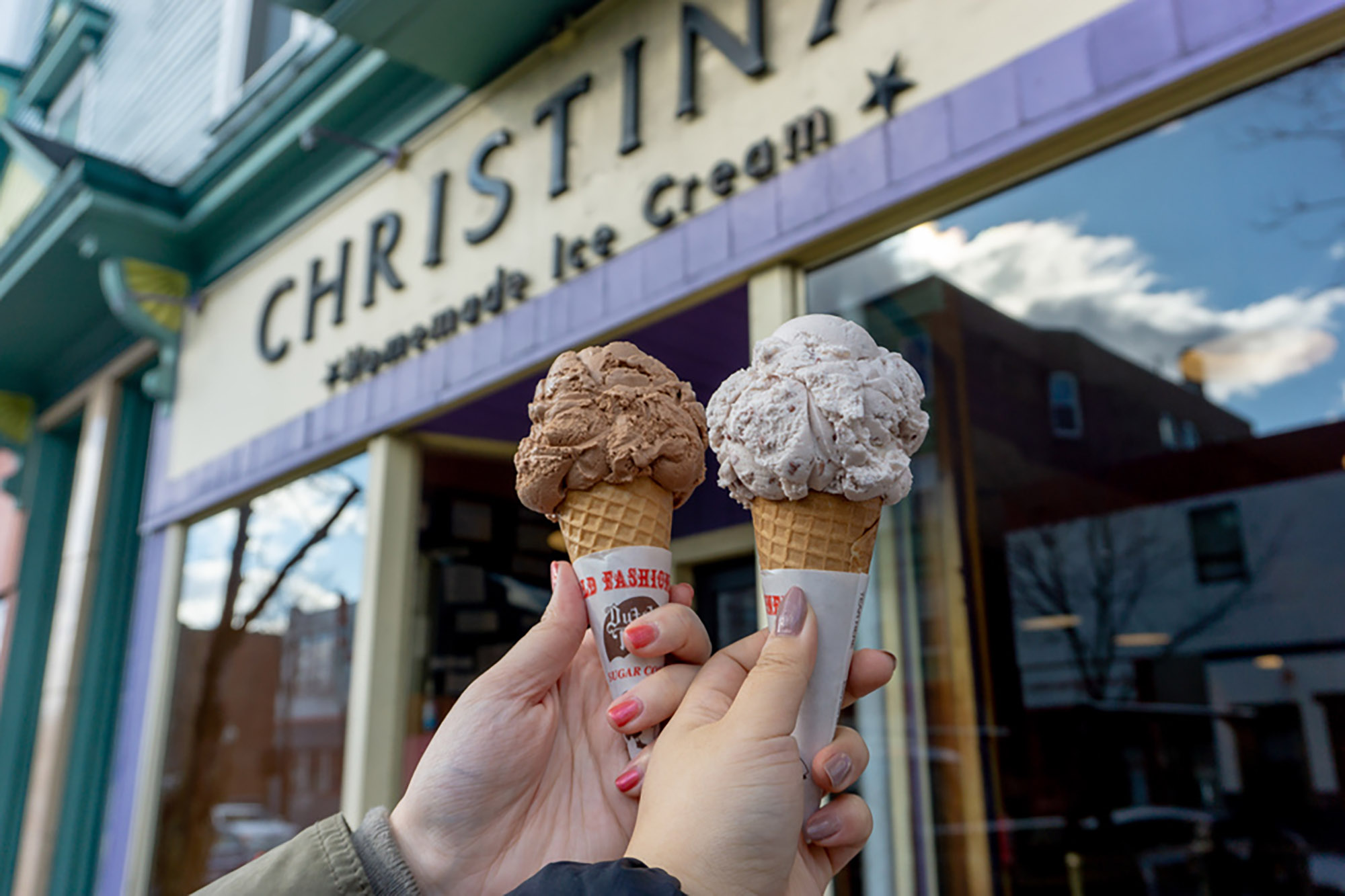 Photo: Two hands hold up cones of ice cream in front of a purple store that reads CHRISTINA ICE CREAM on the front.