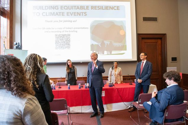 Photo: A meeting room with a powerpoint presentation and four speakers--from left to right, a woman with short curly hair, a woman with straight brown hair, and woman with her hair pulled back, and a man with short hair at the podium. The audience is scattered across the seats available.