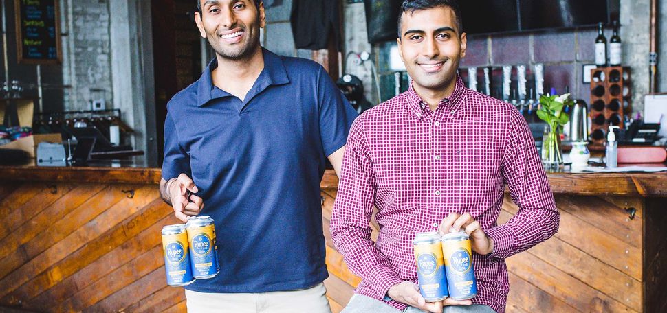 Photo of Sumit Sharma (right, in red) and his brother Vanit Sharma (at left, in blue) posing with four packs of Rupee beer at Dorchester Brewing Company. The brothers are Indian American, have dark hair, and smile as they pose. Sumit sits on a metal stool and Vanit stands. A wooden bar and taps are seen behind them.