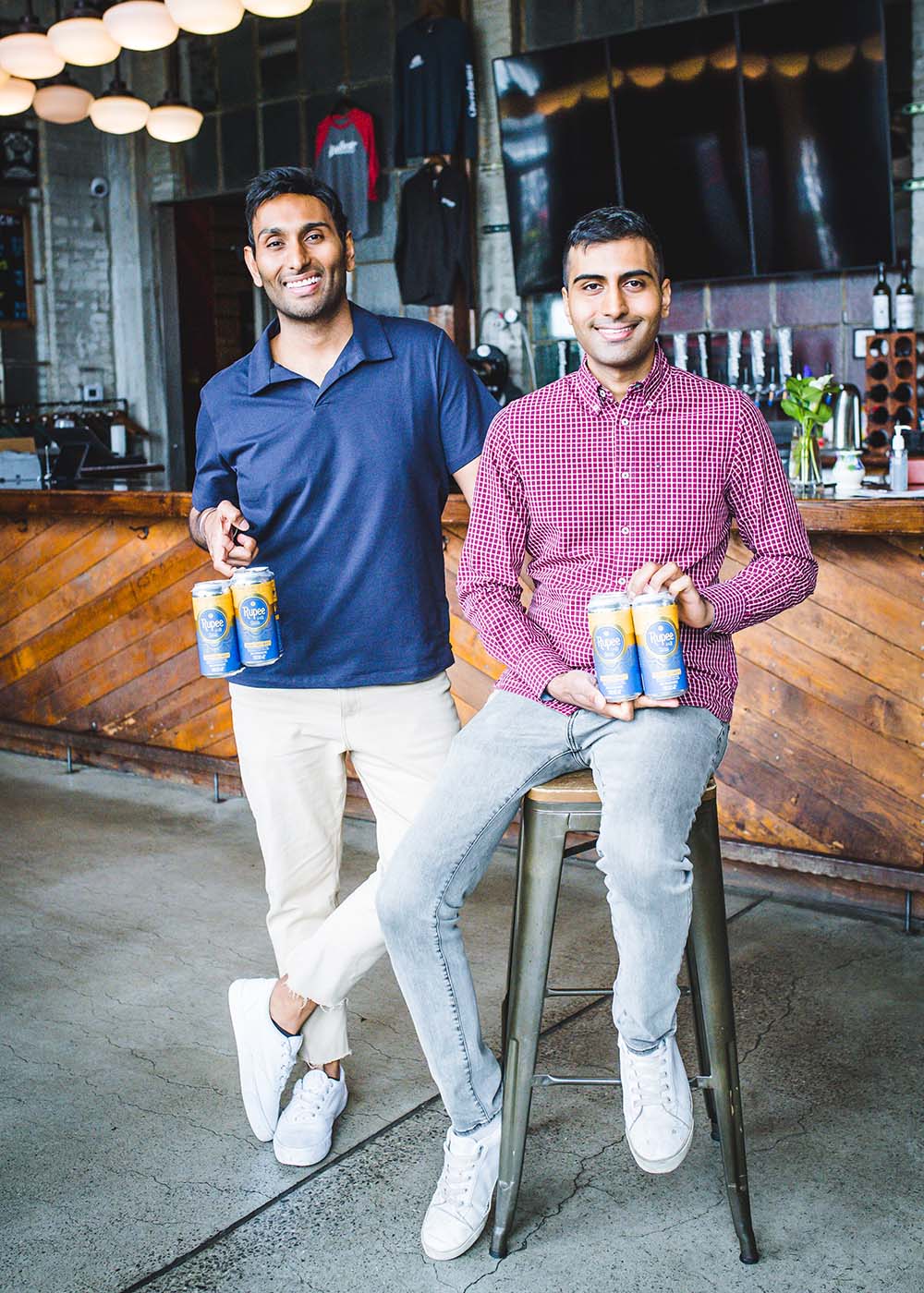 Photo of Sumit Sharma (right, in red) and his brother Vanit Sharma (at left, in blue) posing with four packs of Rupee beer at Dorchester Brewing Company. The brothers are Indian American, have dark hair, and smile as they pose. Sumit sits on a metal stool and Vanit stands. A wooden bar and taps are seen behind them.