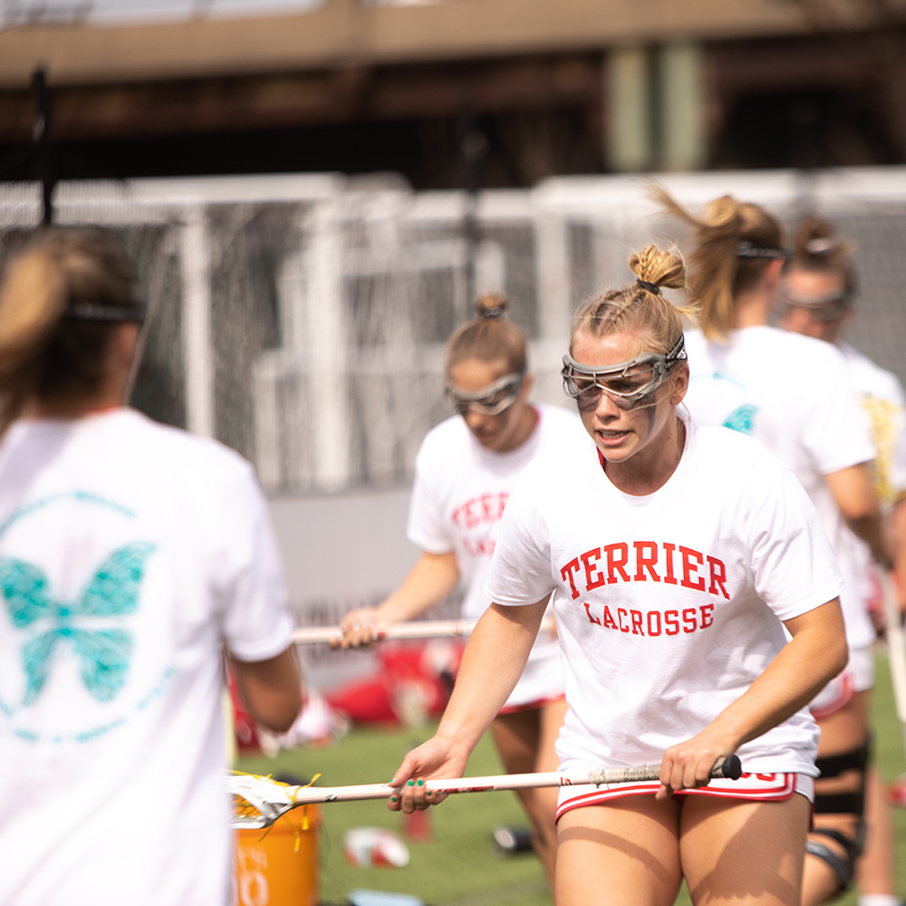 Photo: Action shot of Ellen Monahan as she plays lacrosse. A young white woman wearing a BU white and red lacrosse uniform and goggles looks forward as she practices catching balls with her lacrosse stick.  Another athlete stands in the foreground throwing balls for her to catch.