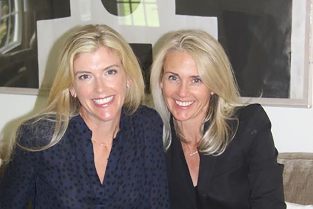 Photo: Two blonde woman, sisters, smile for the camera in dark blouses.