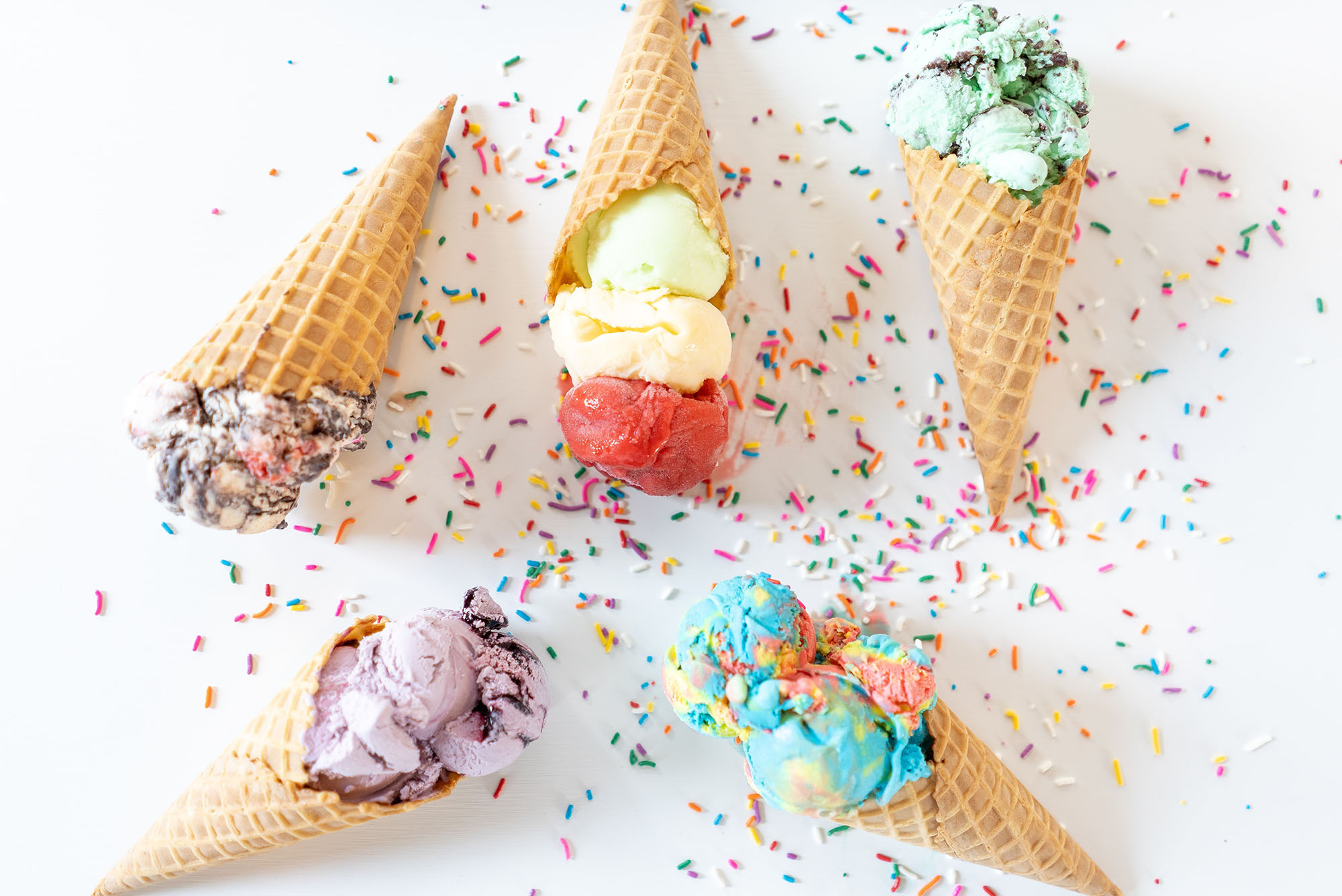 Photo: A white table top with five different ice creams with a waffle cone are scatterd in a flat-lay.