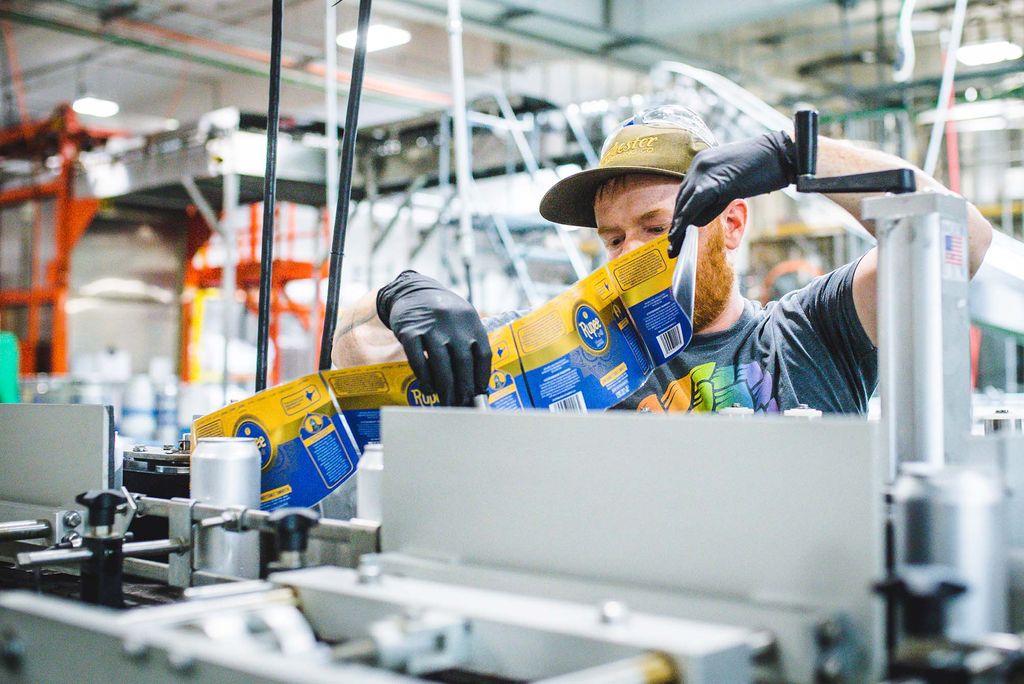 Photo of an employee at Dorchester Brewing Company feeding Rupee beer labels into the wrapping machine. The employee is White with a reddish beard; he wears a baseball hat and black gloves. Brewery machinery is seen in the foreground and background