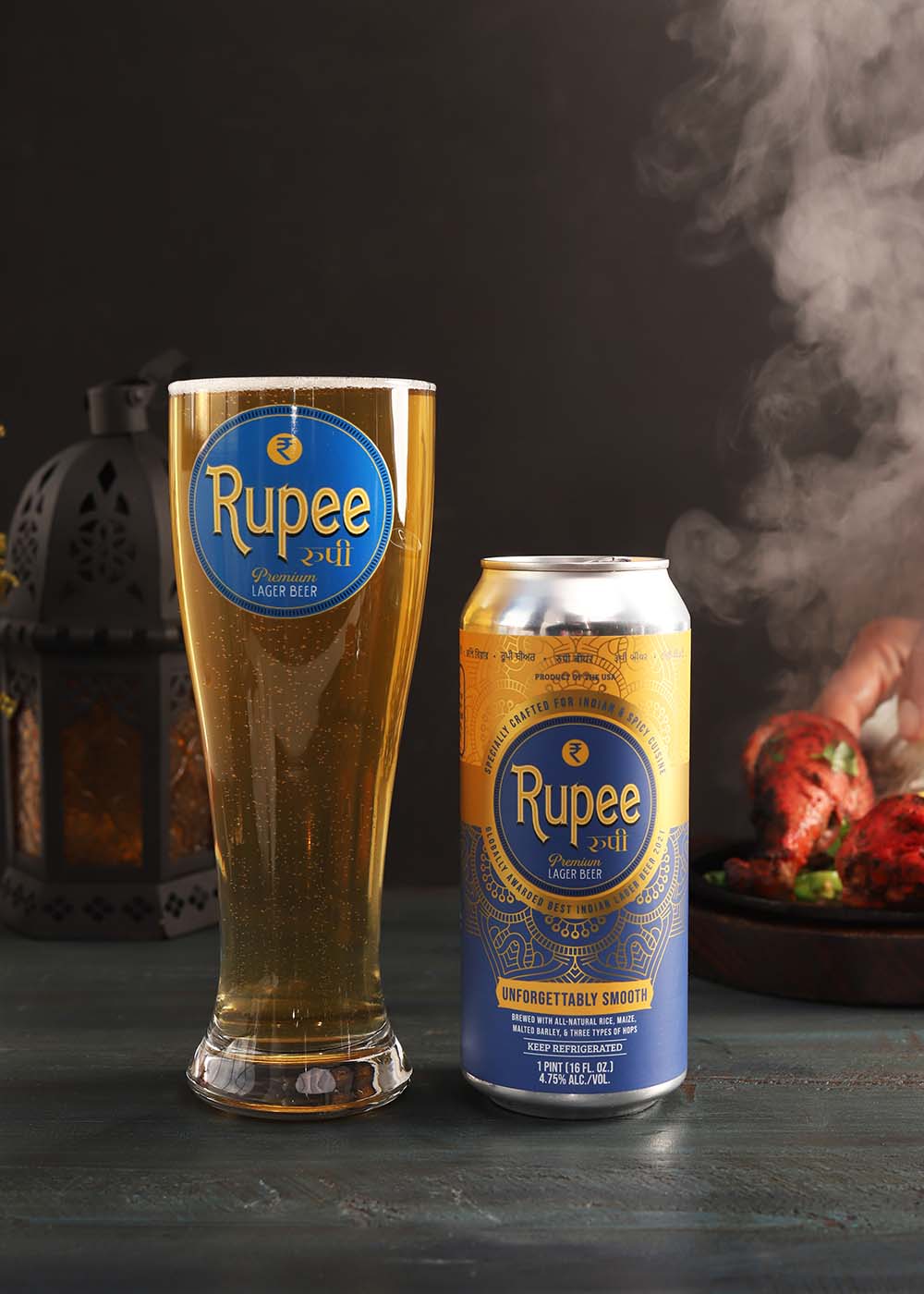 Photo of a tall glass of beer next to a can of Rupee beer. The glass and can both have Rupee's gold and blue logo. In the background, steaming Indian food can be seen. The can and glass are brightly lit, the background is mostly black.