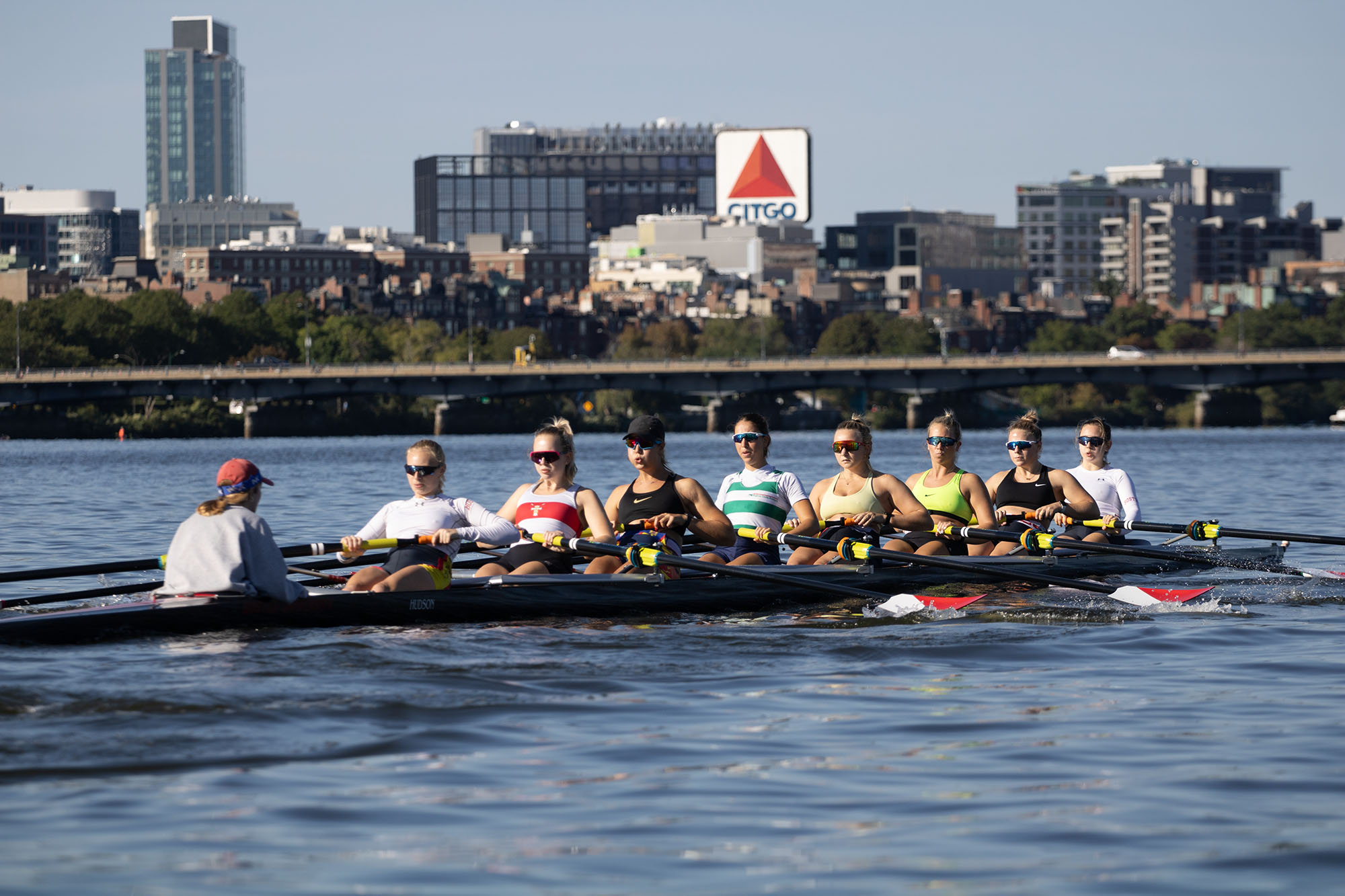 Photo: A boat full of women rowers are shown rowing down the Charles River in Boston on a sunny day. The rowers wear sports bras and sunglasses as the Boston city skyline can be seen in the background.