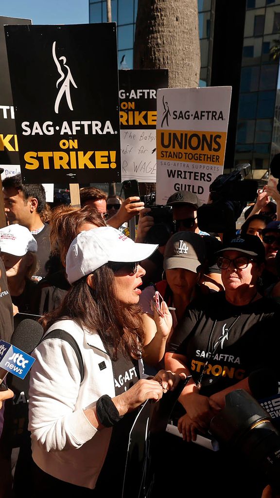 Photo: Fran Drescher, President of SAG, is swarmed by news outlets as she heads to a press call. She is wearing all white and stands in the middle while cameras and microphones surround her.