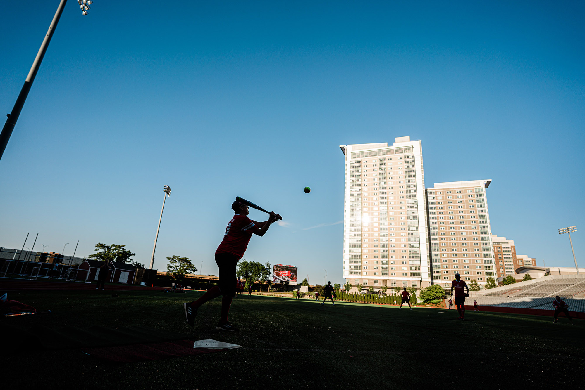 Photo: A batter swings to hit a ball on the field with buildings in the background.